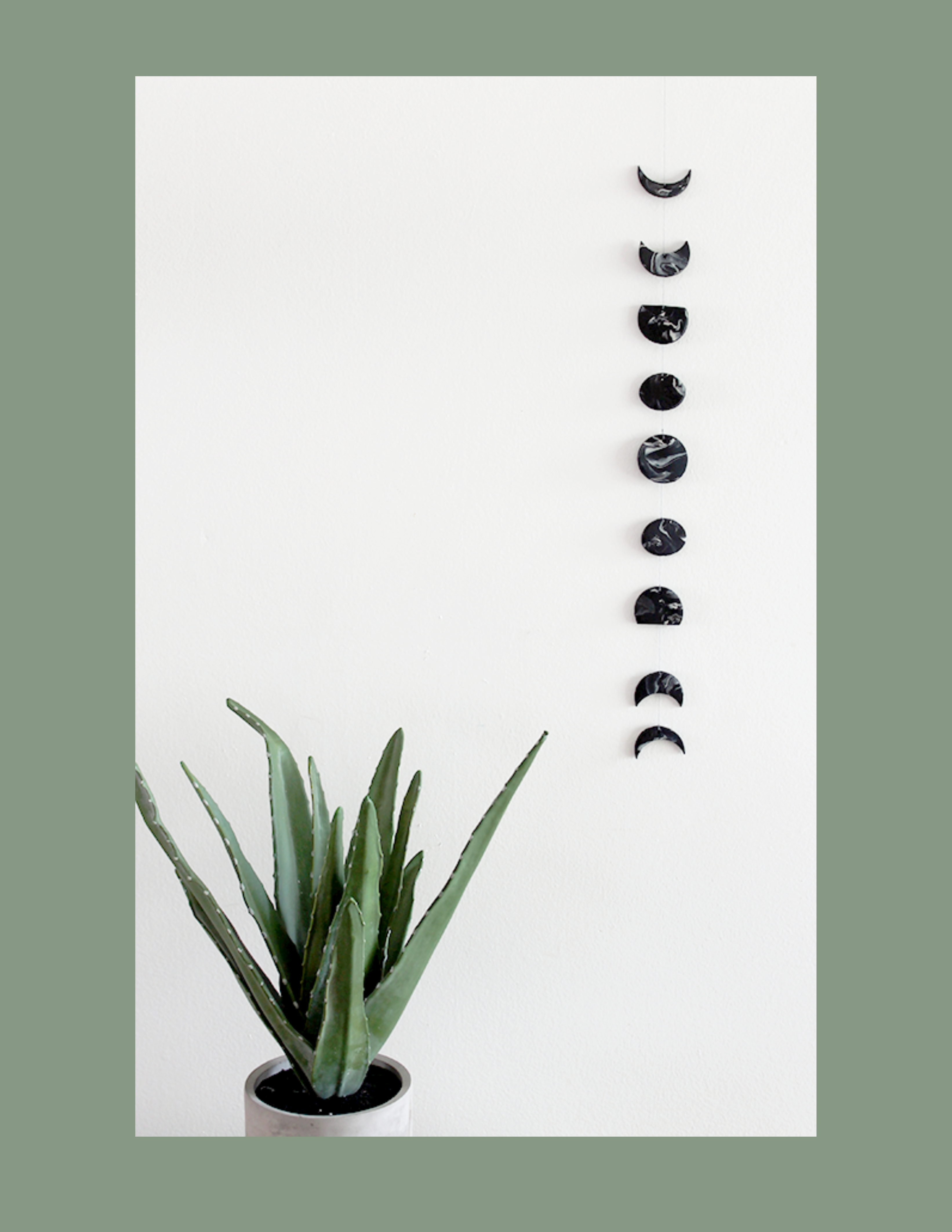 Moon phase hanging art in black against a white background with green plant in pot on the left side. 
