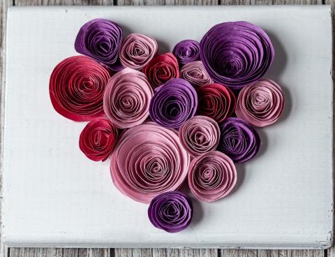 Multicolored rolled circles in the shape of a heart against a white background with some wood planks showing. 