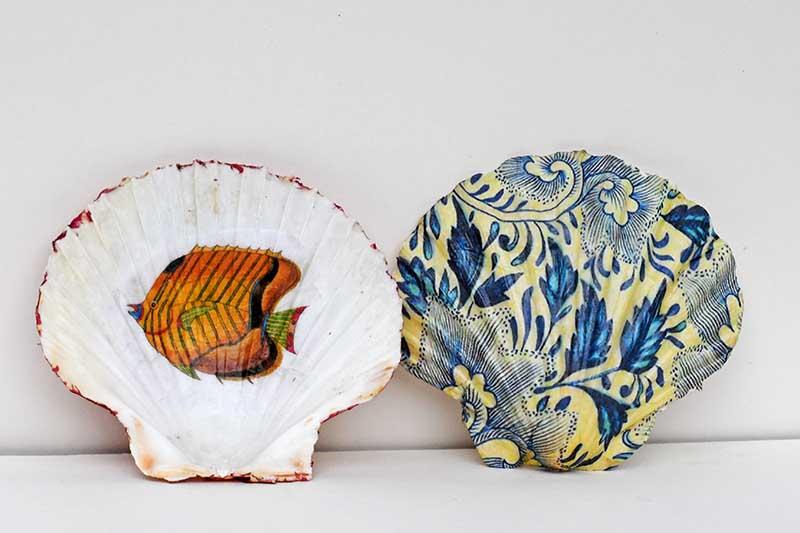 Two decoupaged seashells. One with a fish on it and one with a floral background. Both shells are against a white background. 