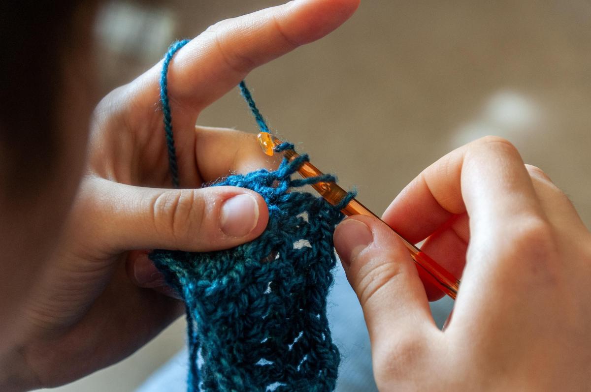 Hands crocheting a blue yarn against a wood floor. 